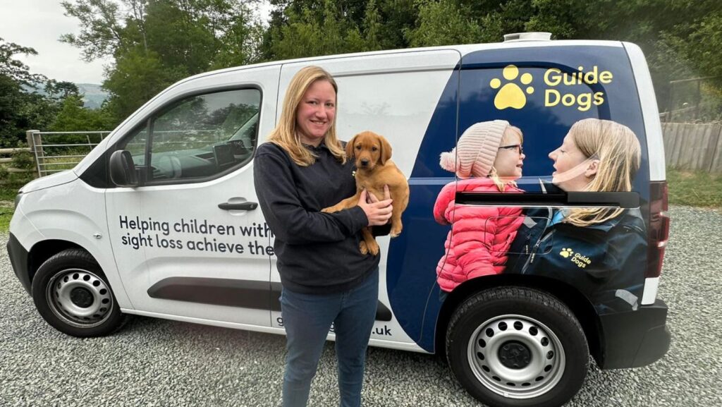 labrador puppy in arms of woman next to guide dogs for the blind vehicle